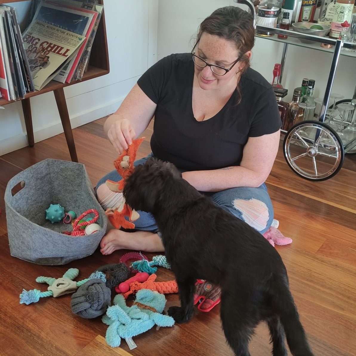 A woman in a black shirt sits on the ground and plays with a black dog.
